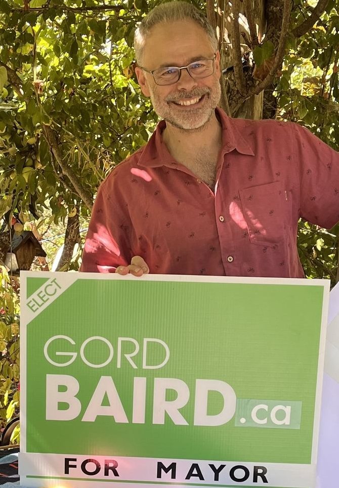 Gord Baird smiling under the kiwi vines at Eco-Sense, holding a green campaign sign reading Elect Gord Baird.ca for Mayor