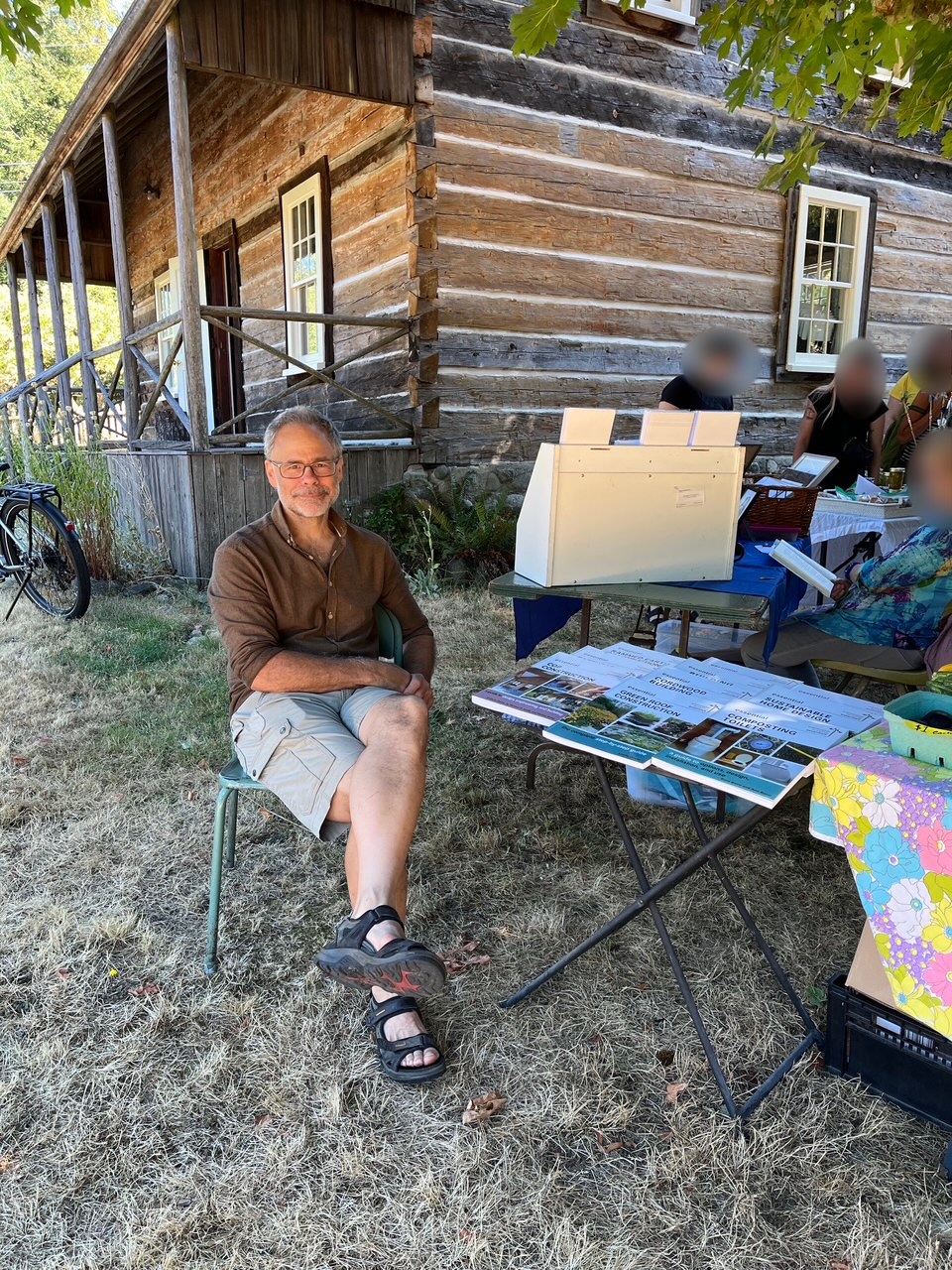 Gord Baird at a Highlands community market table beside the Essential series building and sustainability books, in front of the historic Caleb Pike log building